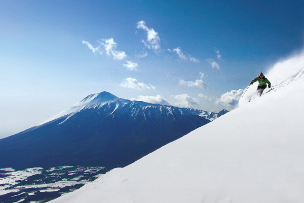 青空と山が見える雪の斜面をスキーで滑走している写真。